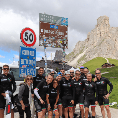 Group of cyclists from Velo Tours standing together smiling under the summit sign of the Giau Pass celebrating their ride up the mountain